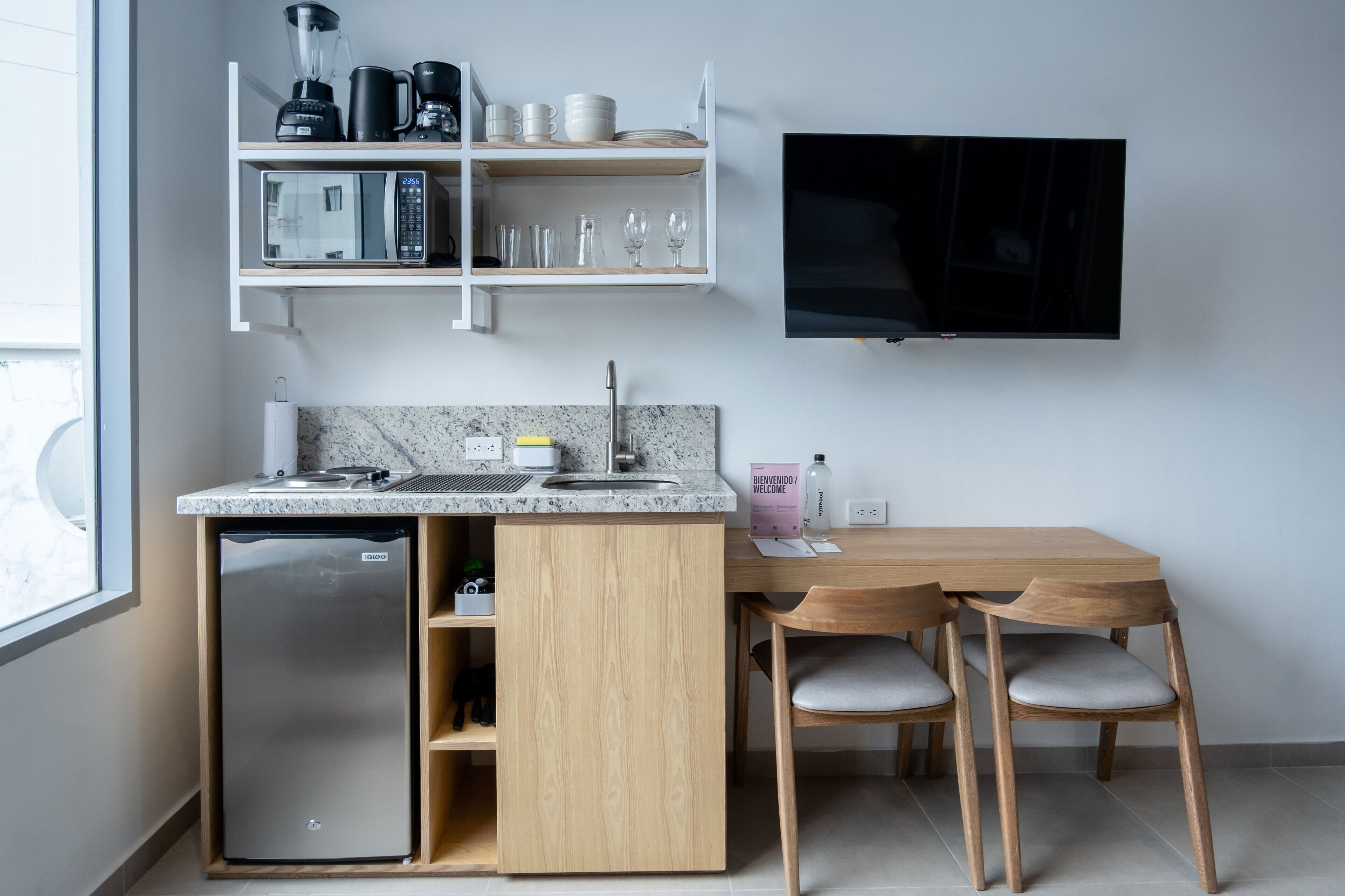 Cocina compacta con refrigerador empotrado, microondas y cafetera en la alacena. Hay una mesa de comedor de madera con dos sillas y un televisor de pantalla plana montado en la pared.
