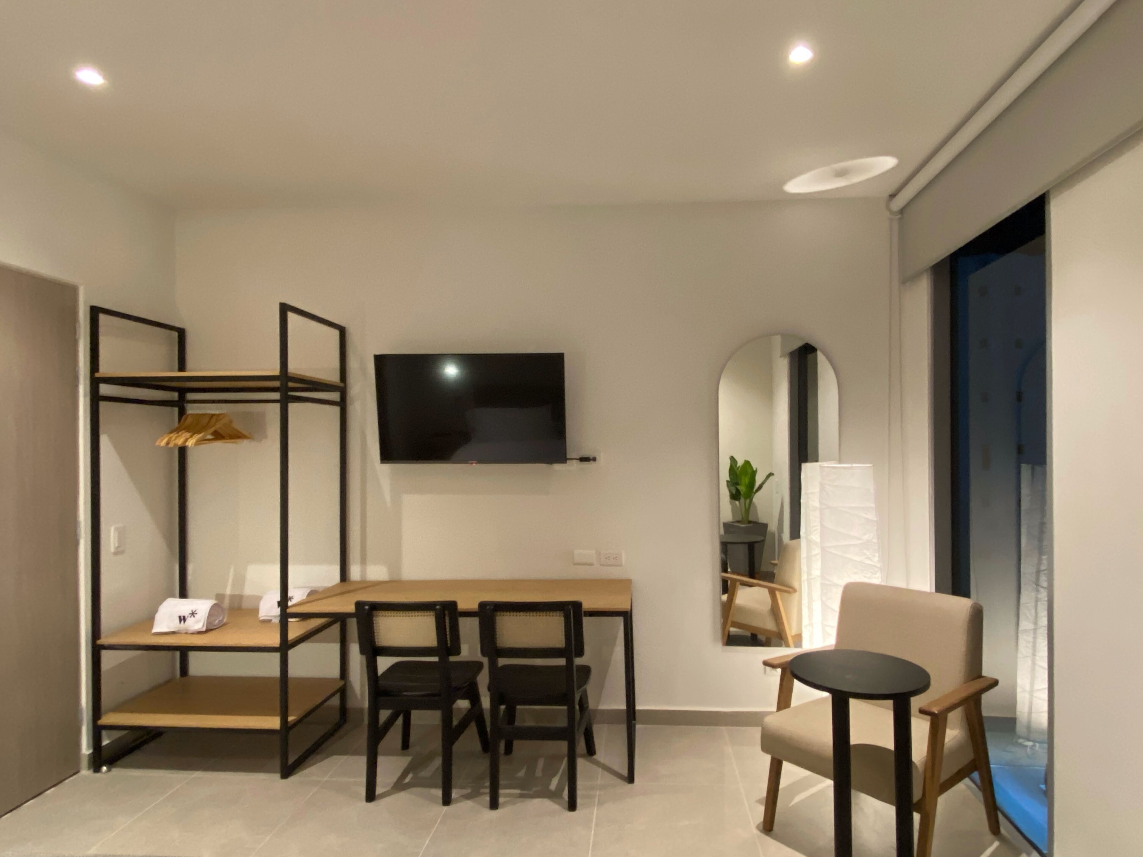 A modern hotel room with a wooden dining table, two black chairs, a TV mounted on the wall, a mirror, and a window with a view.