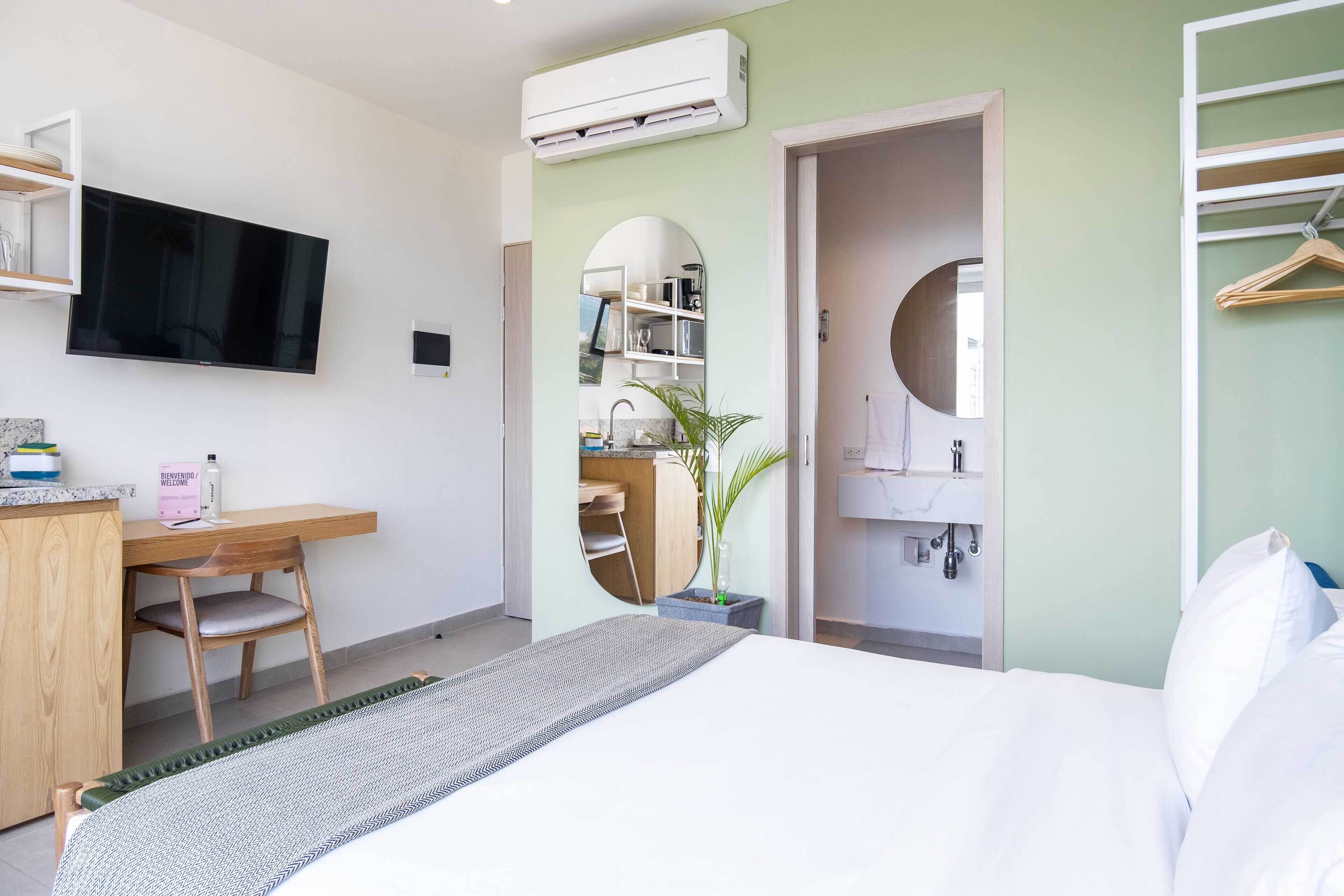 A modern hotel room with a bed, a desk with a chair, a television, a mirror, and a wardrobe.