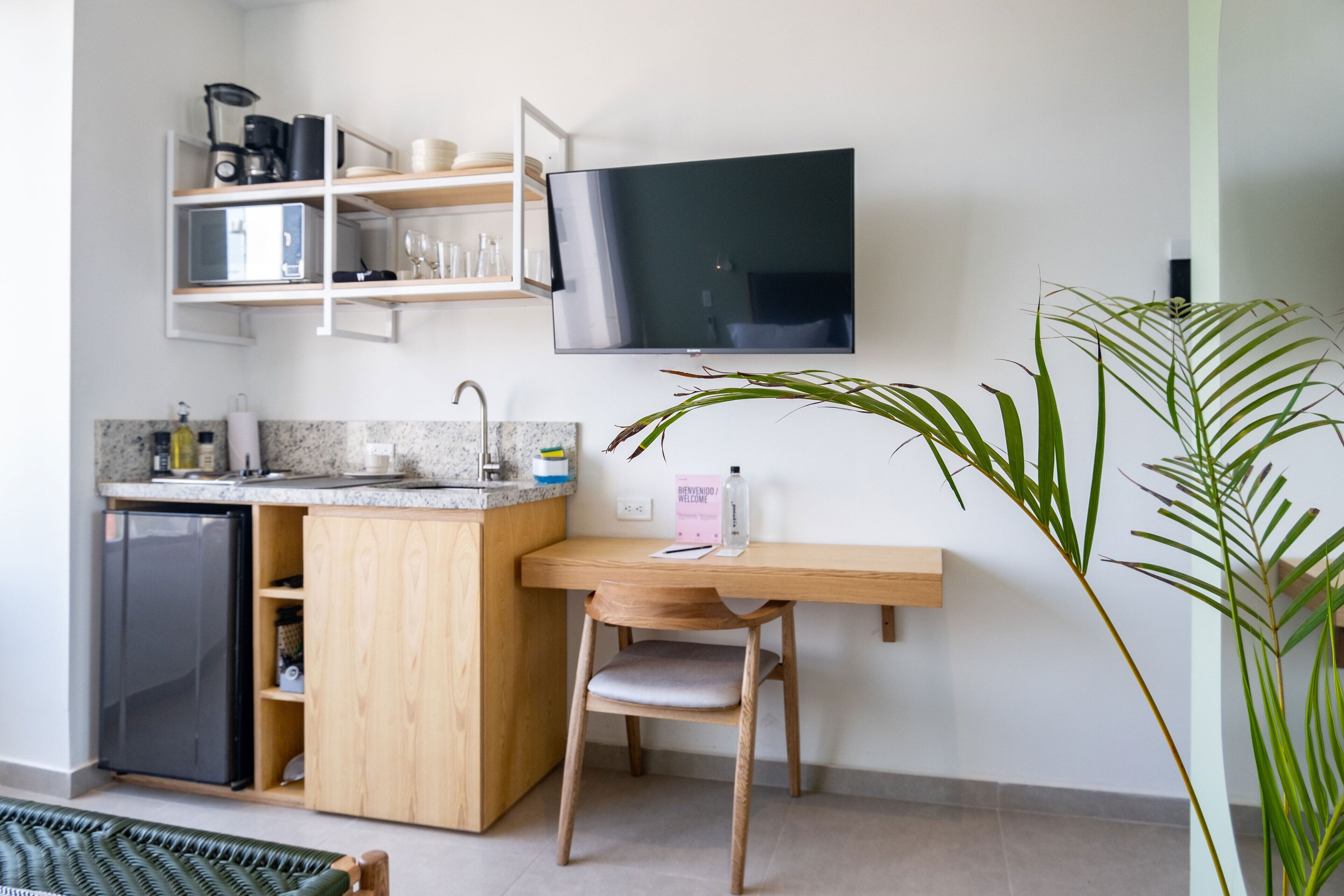 A compact kitchen with a built-in fridge, a wooden dining table, a TV mounted on the wall, and a potted plant.