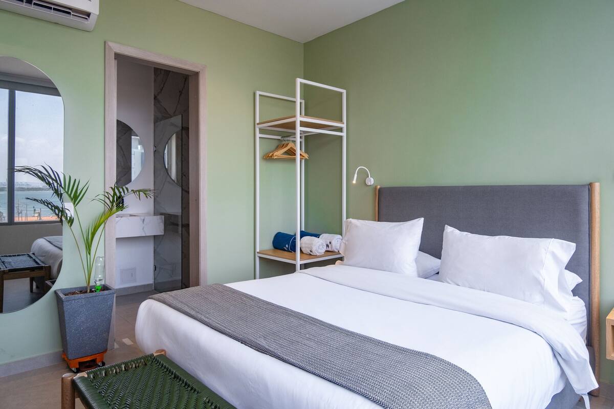 Ein ordentlich eingerichtetes Hotelzimmer mit Bett, Nachttisch, Spiegel, einer Pflanze und einem Kleiderschrank mit Kleidung.