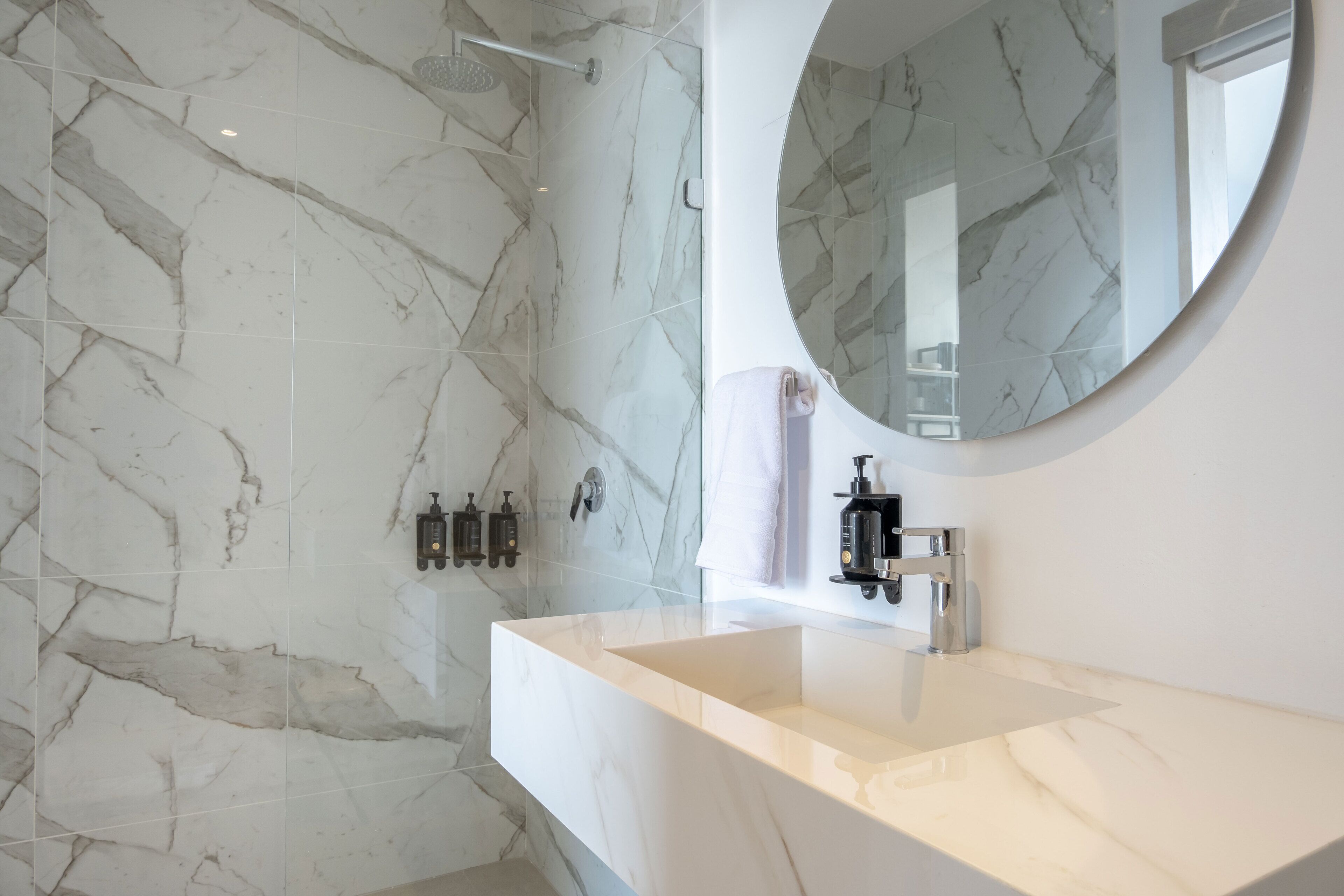 A modern bathroom with a marble wall, a large round mirror, and a white sink.