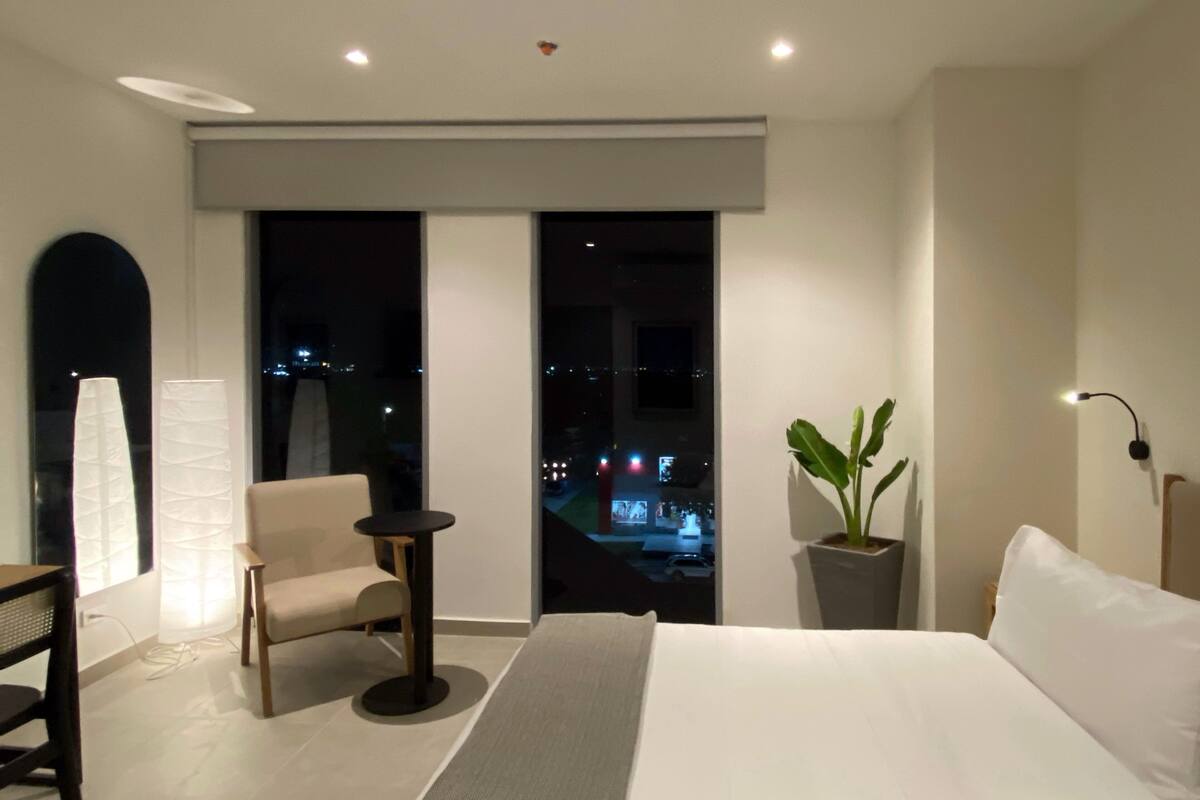 Ein modernes Hotelzimmer mit einem großen Bett, einer Sitzecke mit Sessel und kleinem Tisch, einer Topfpflanze und Blick auf die beleuchtete Stadt bei Nacht.
