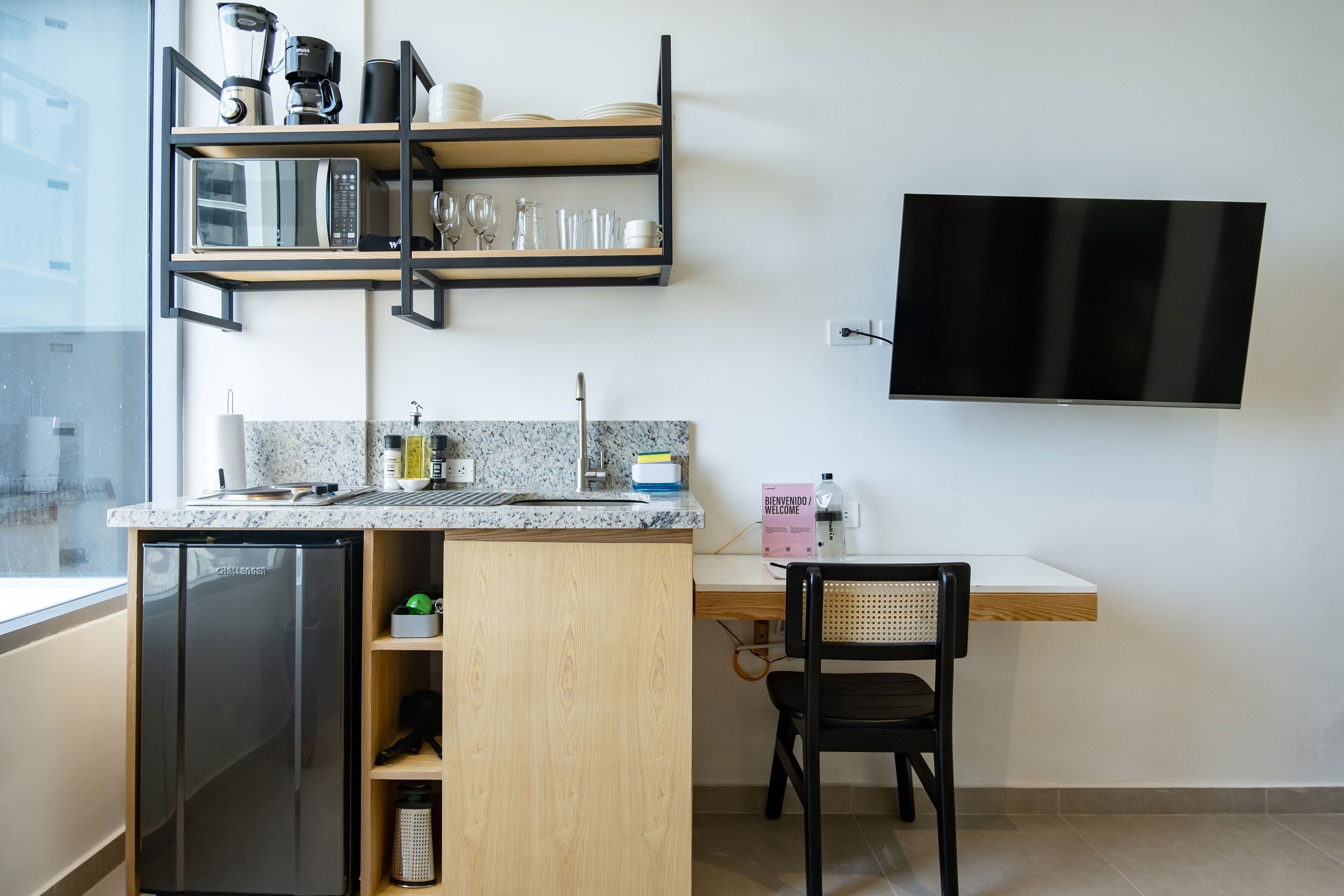 Une cuisine compacte équipée d’un lave-vaisselle encastré, d’un four à micro-ondes et d’une machine à café placés sur les étagères. On y trouve un petit évier avec un robinet, un plan de travail avec un distributeur de savon et une chaise. Un téléviseur à écran plat est fixé au mur.