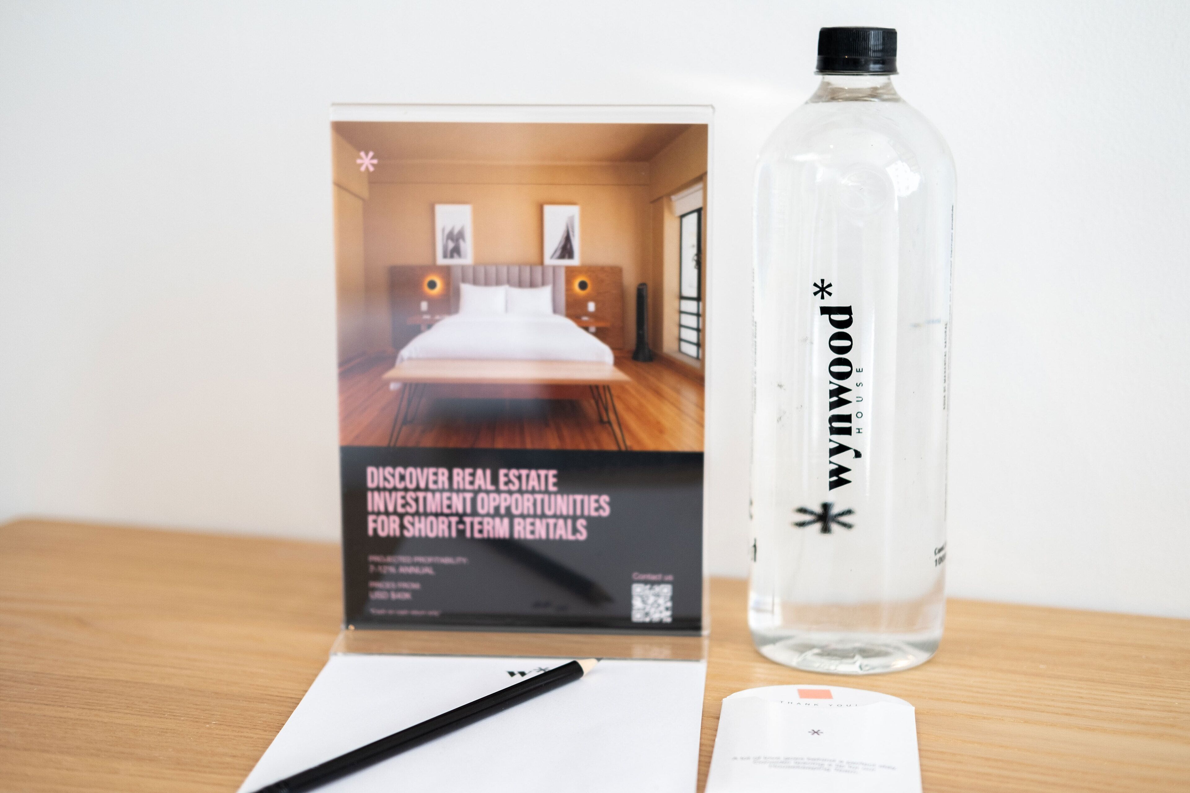 A brochure for real estate investment opportunities for short-term rentals, a clear plastic bottle with a label, a black pen, and a thank you card on a wooden surface.