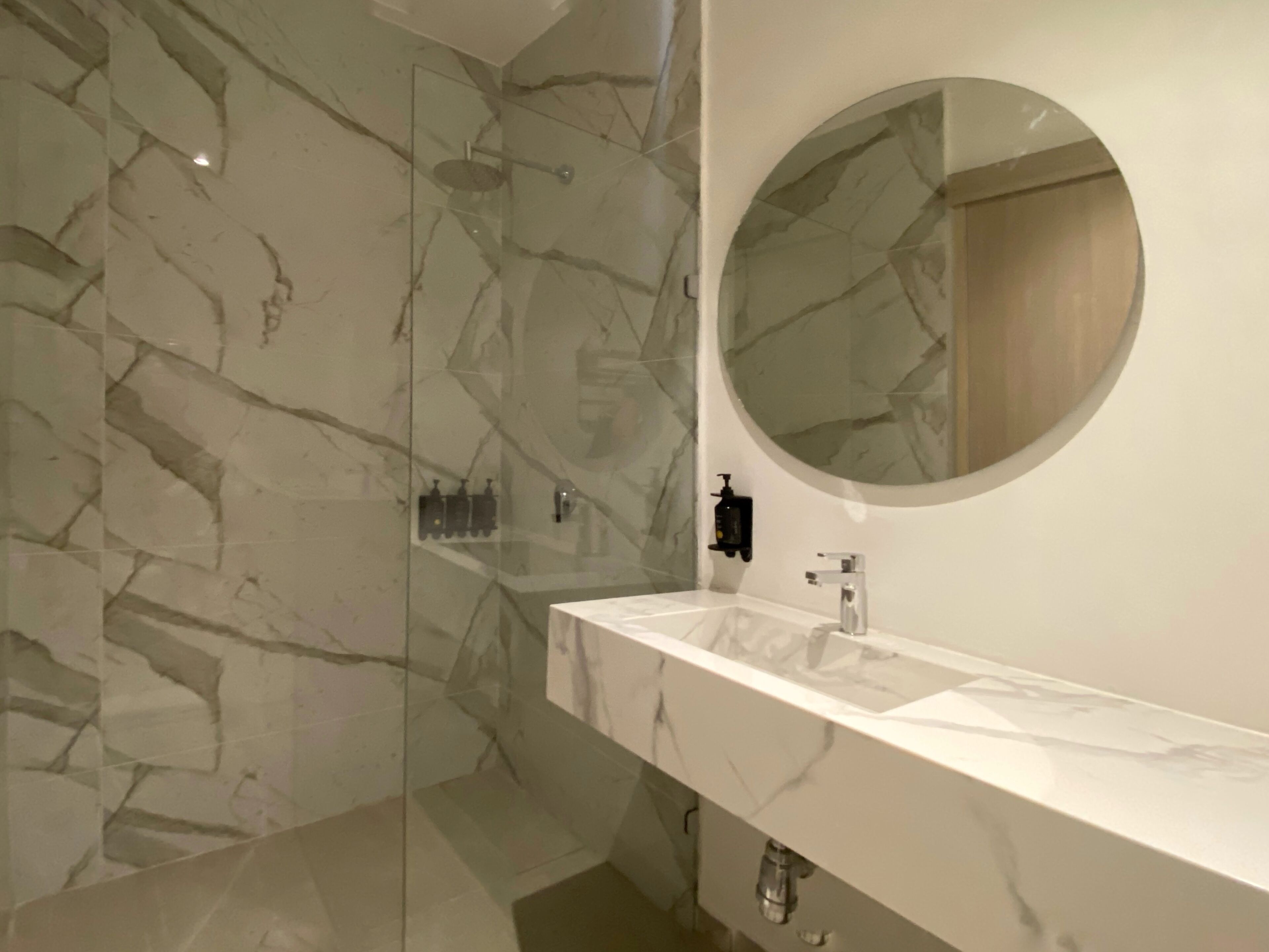 A modern bathroom with a marble vanity, a glass-enclosed shower, and a round mirror.