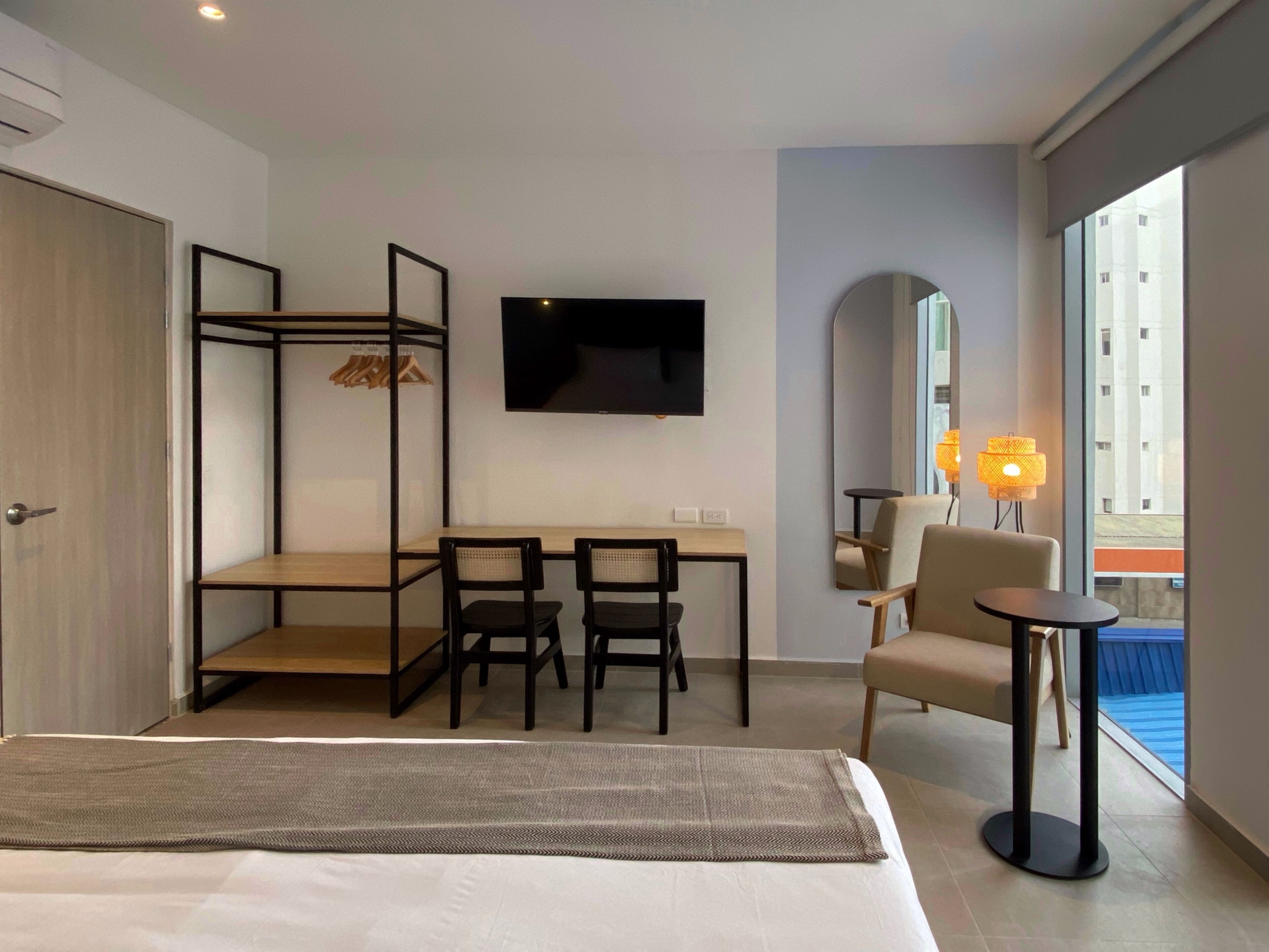 A hotel room with a bed, a desk with two chairs, a TV, a mirror, and a window with a view of buildings.