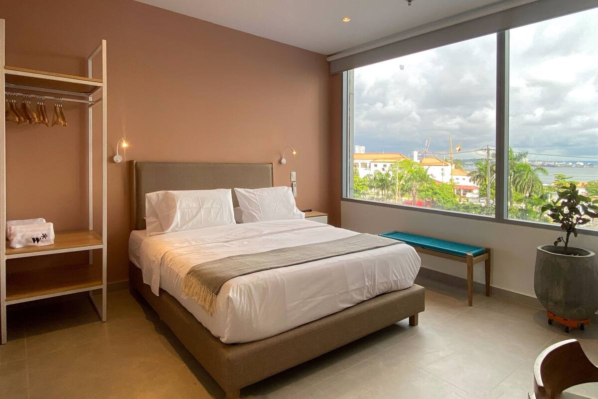 Ein modernes Hotelzimmer mit einem großen Bett, einem Kleiderschrank, einer Bank und Blick auf die Stadt.