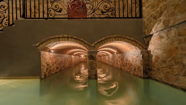 A stone-walled, arched tunnel with a reflective water surface and a central column.