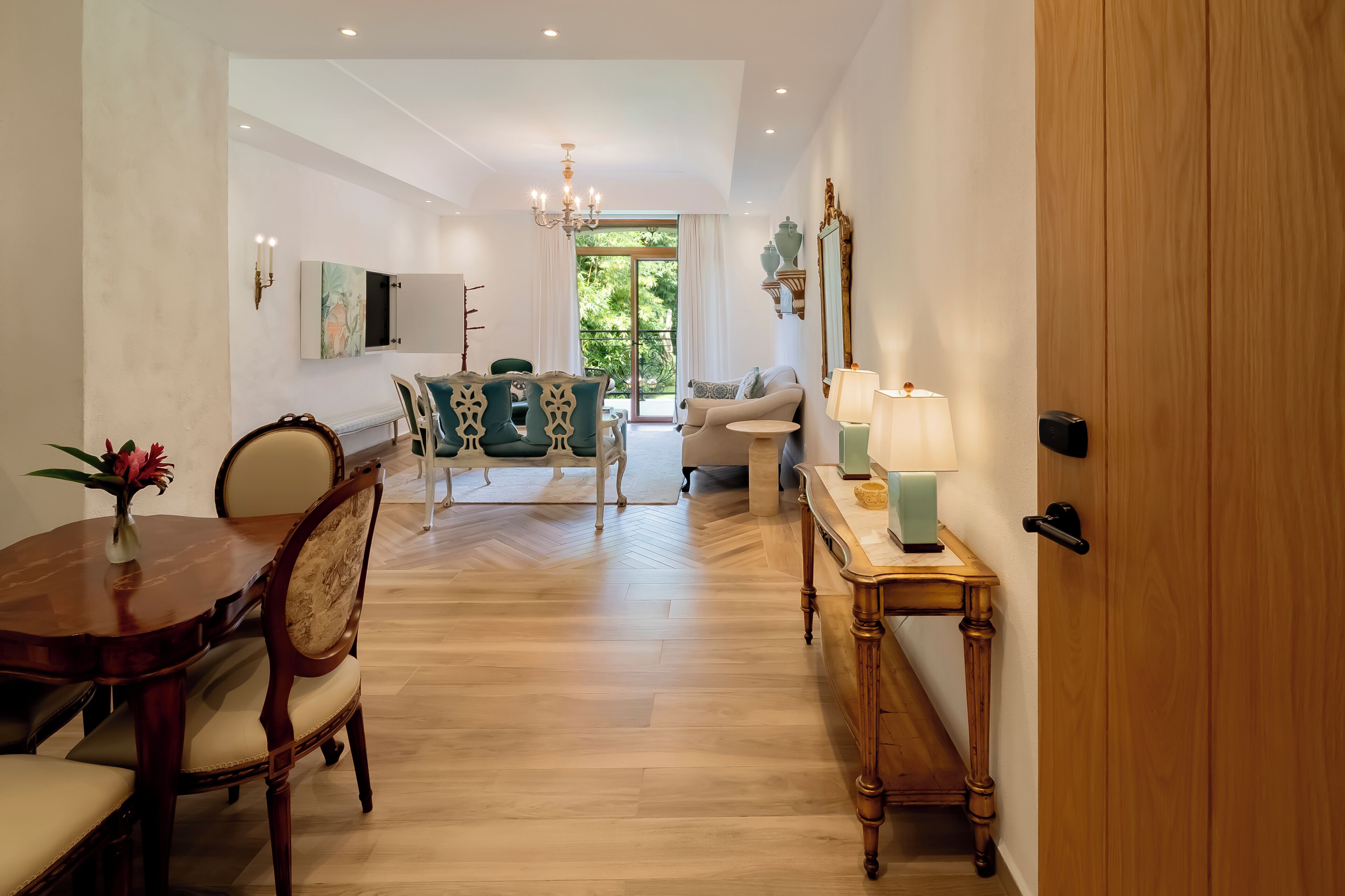 A spacious living area with a wooden floor, a dining table with chairs, a sofa, and a side table with a lamp.