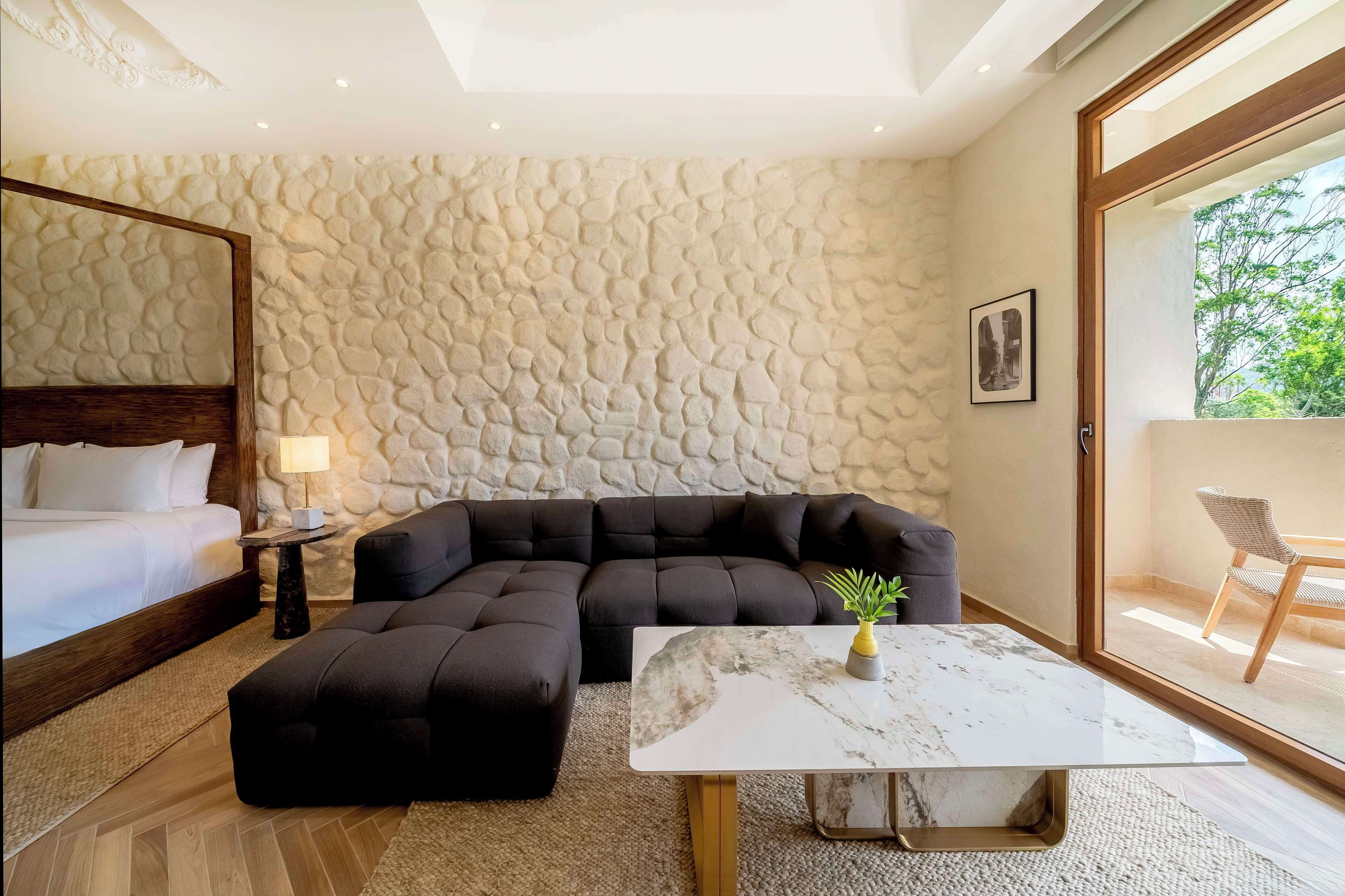 A modern bedroom with a stone wall, a large bed, a sectional sofa, and a marble coffee table.