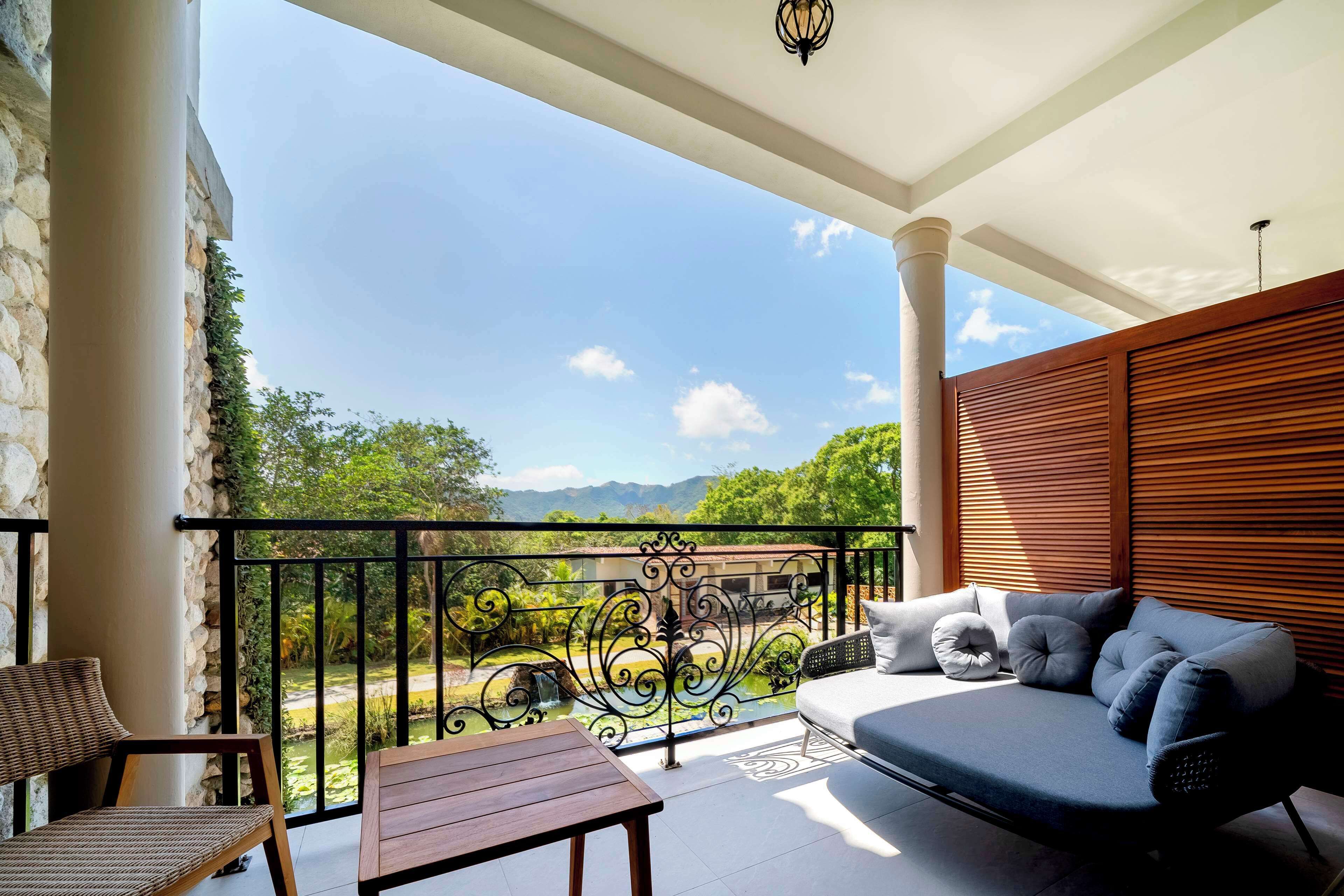 A balcony with a sofa, a table, and a chair, overlooking a garden and mountains.