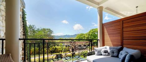 A balcony with a sofa, a table, and a chair, overlooking a garden and mountains.