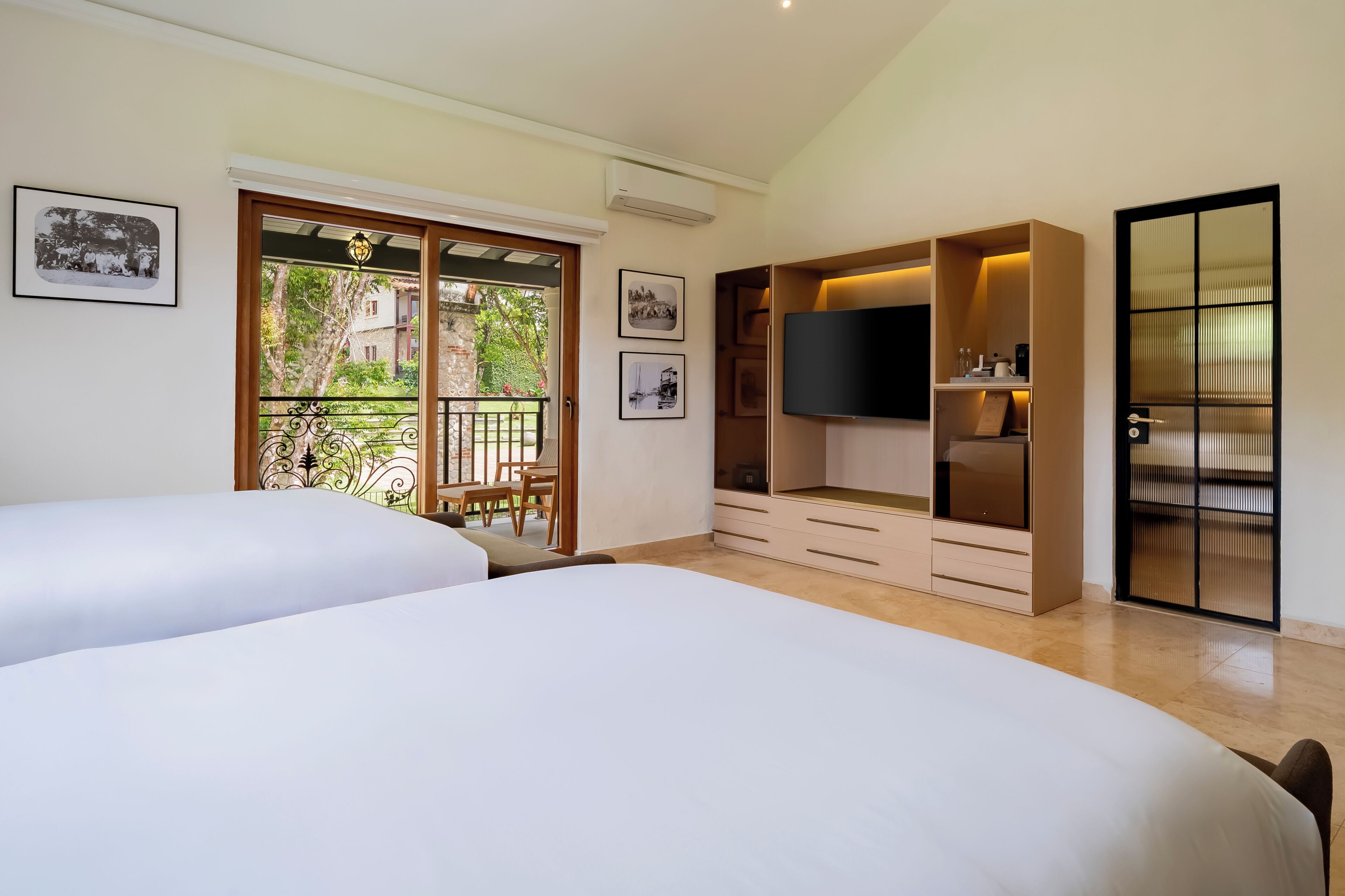 Een slaapkamer met twee bedden, een televisie, een kledingkast en een balkon met uitzicht op groen.