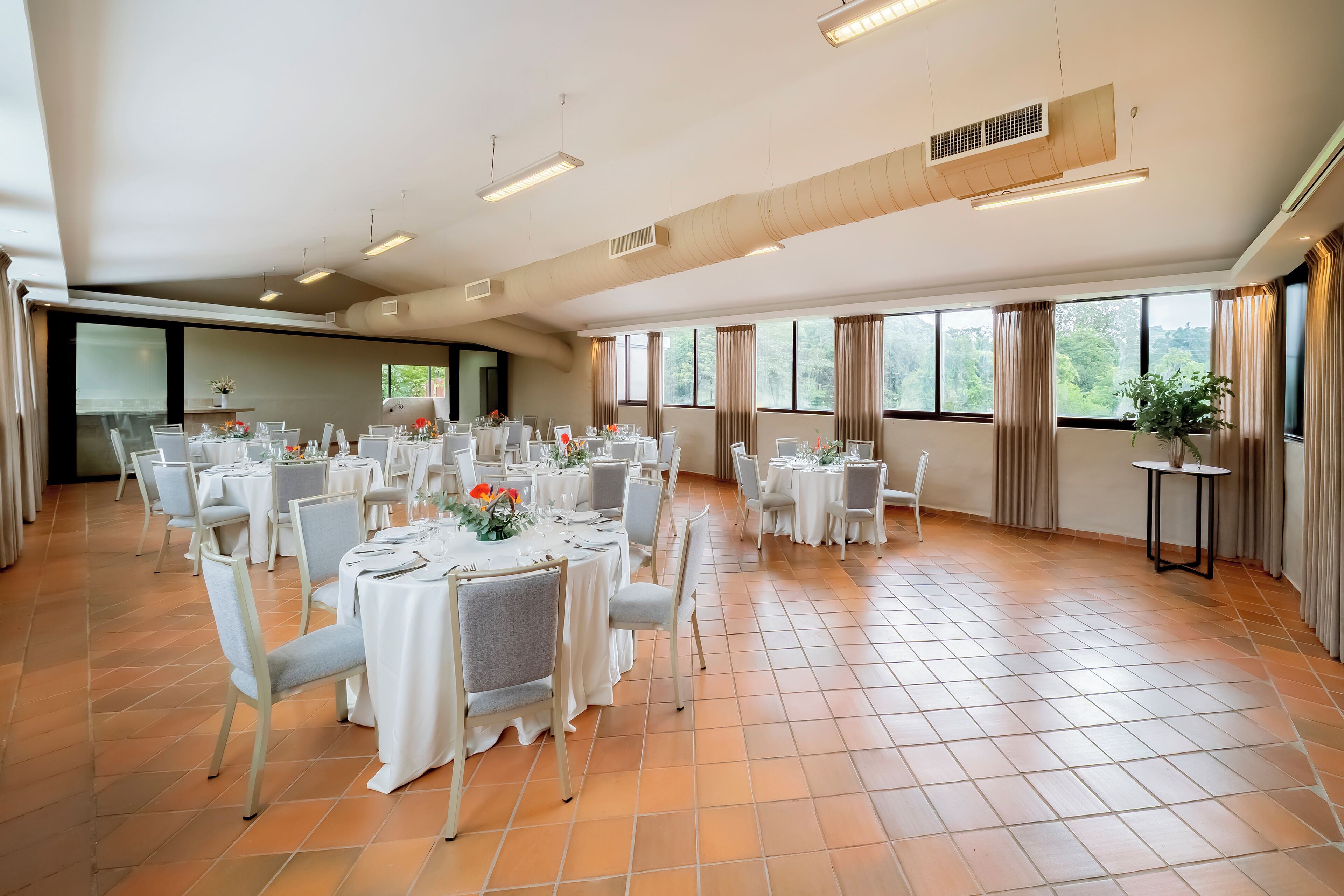 A spacious banquet hall with multiple round tables set for dining, large windows with curtains, and a tiled floor.