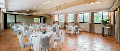 A spacious banquet hall with multiple round tables set for dining, large windows with curtains, and a tiled floor.