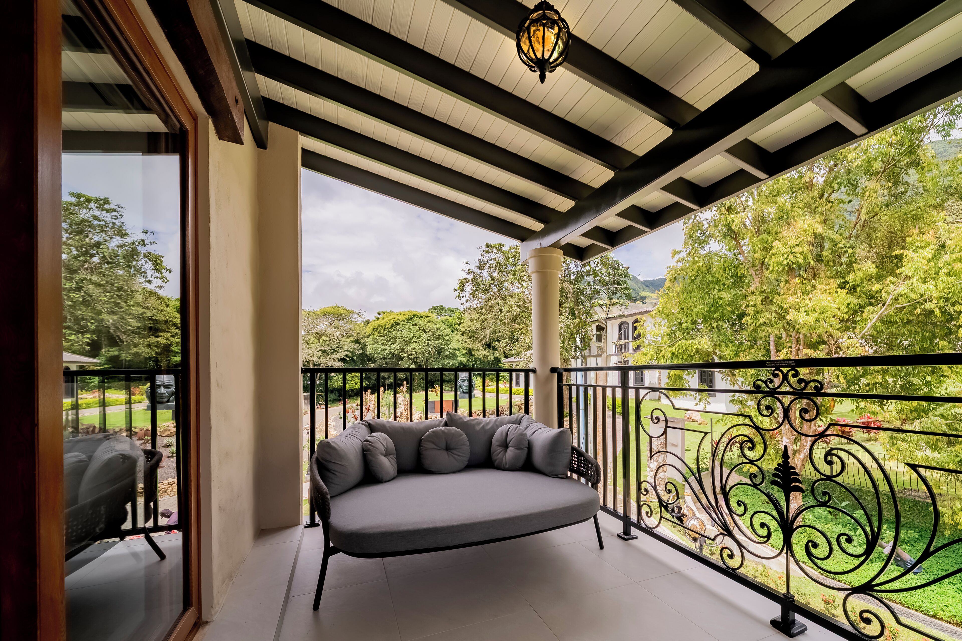 A covered balcony with a grey sofa and a view of a garden and building.
