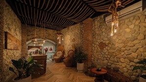 A cozy interior with a stone wall, wooden floor, and a rustic ceiling. There are potted plants and a seating area with a curved bench.