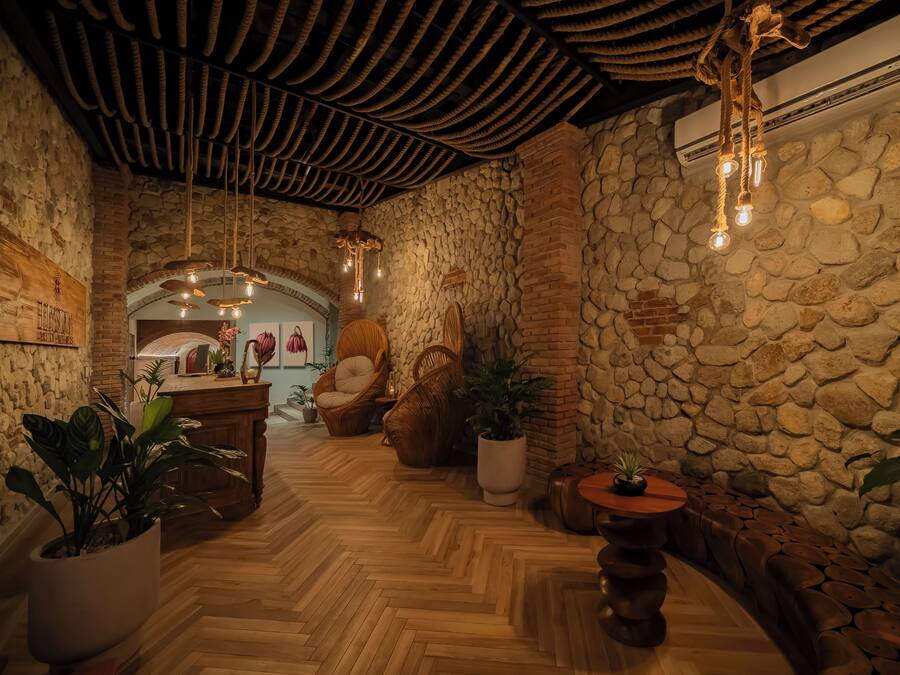 A cozy interior with a stone wall, wooden floor, and a rustic ceiling. There are potted plants and a seating area with a curved bench.
