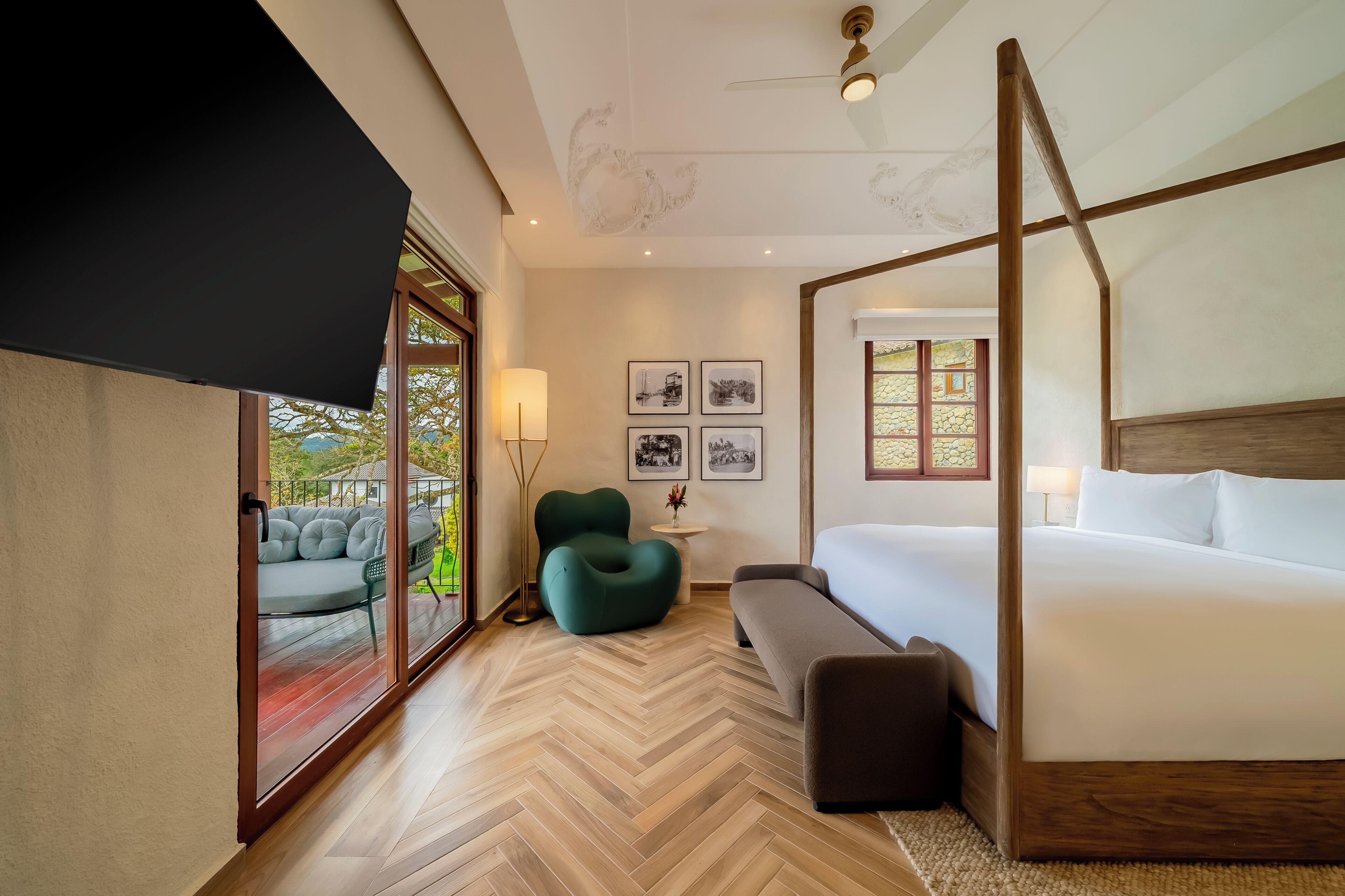 A modern bedroom with a four-poster bed, a herringbone wood floor, a green armchair, and a view through large glass doors.