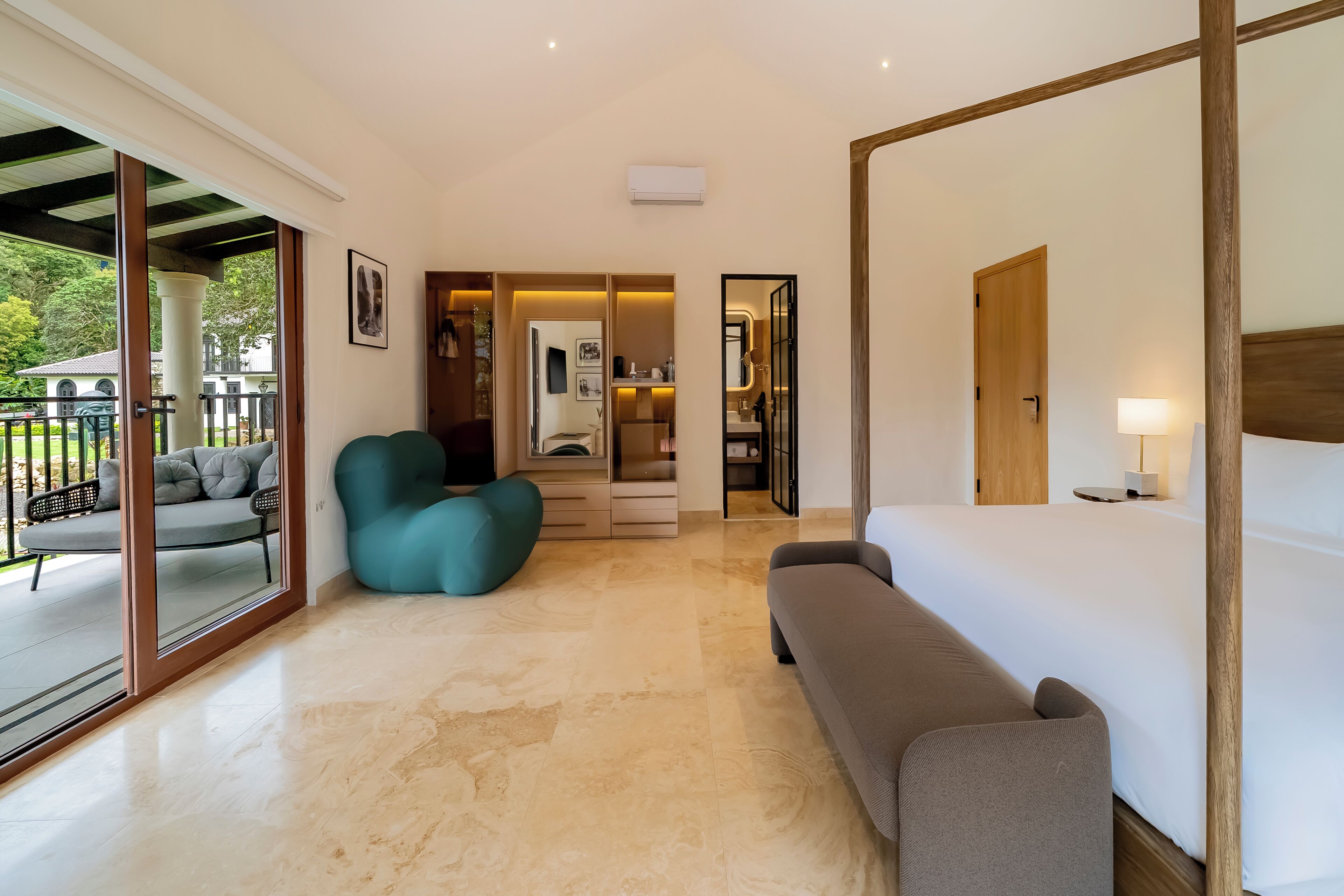 A spacious hotel room with a large bed, a sofa, a teal armchair, and a view of the outdoors through a sliding glass door.