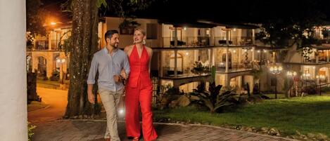 A couple walking on a paved pathway at night, with a building featuring balconies and outdoor lighting in the background.