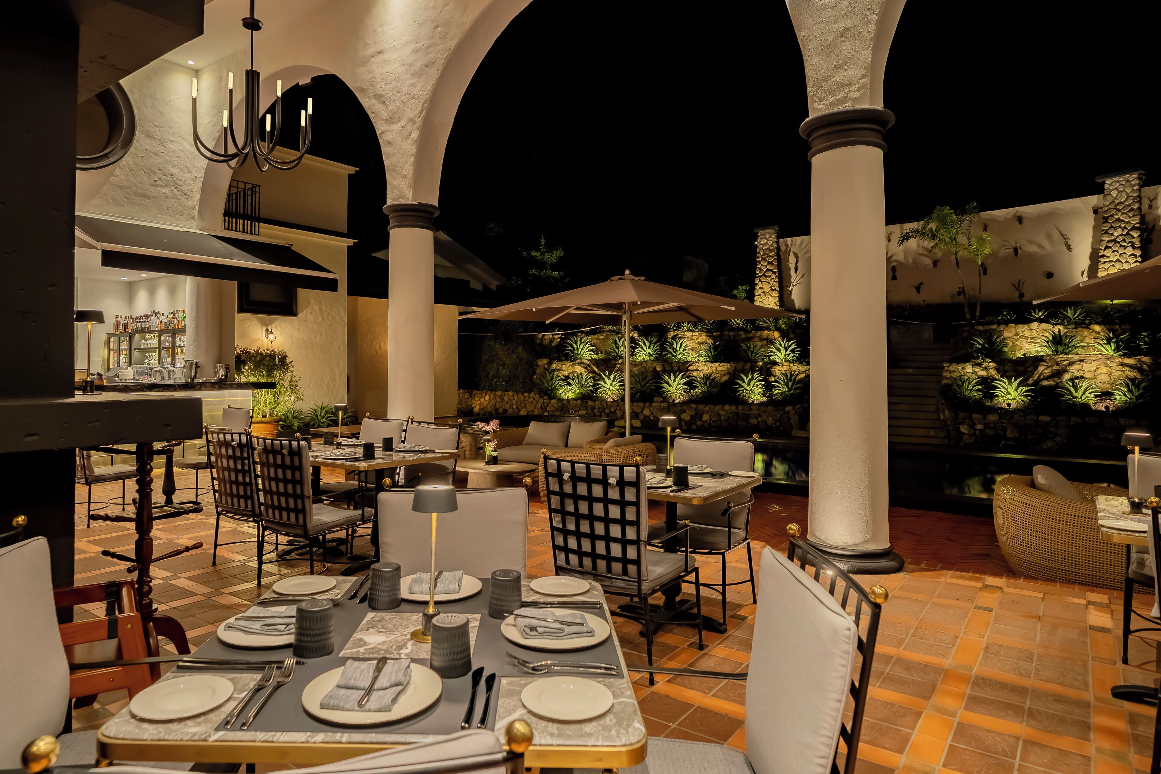 An outdoor dining area with tables set for two, surrounded by columns and potted plants.