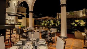 An outdoor dining area with tables set for two, surrounded by columns and potted plants.
