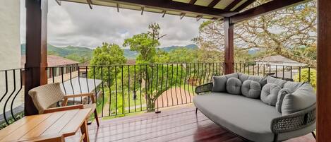A covered outdoor area with a wooden table and chairs, a cushioned sofa, and a view of greenery and mountains.
