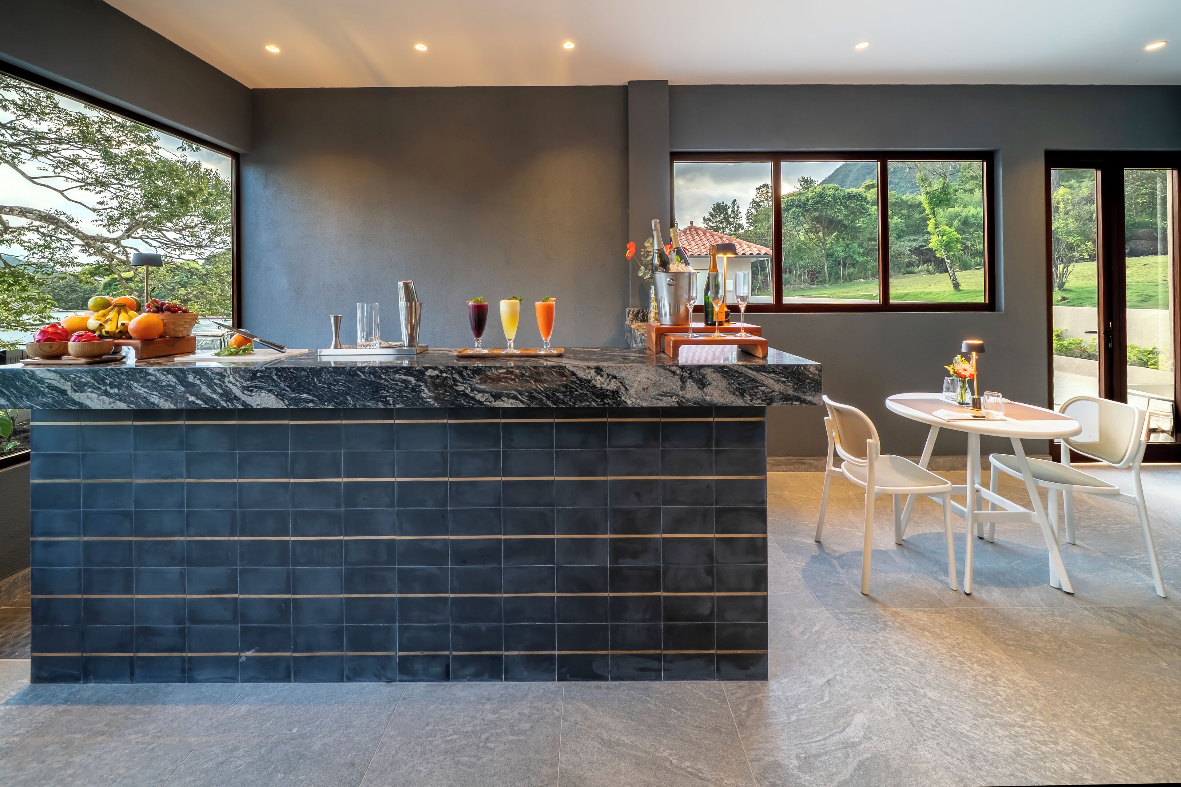 A modern kitchen with a marble island, a dining area with white chairs, and large windows offering a view of greenery and a house.