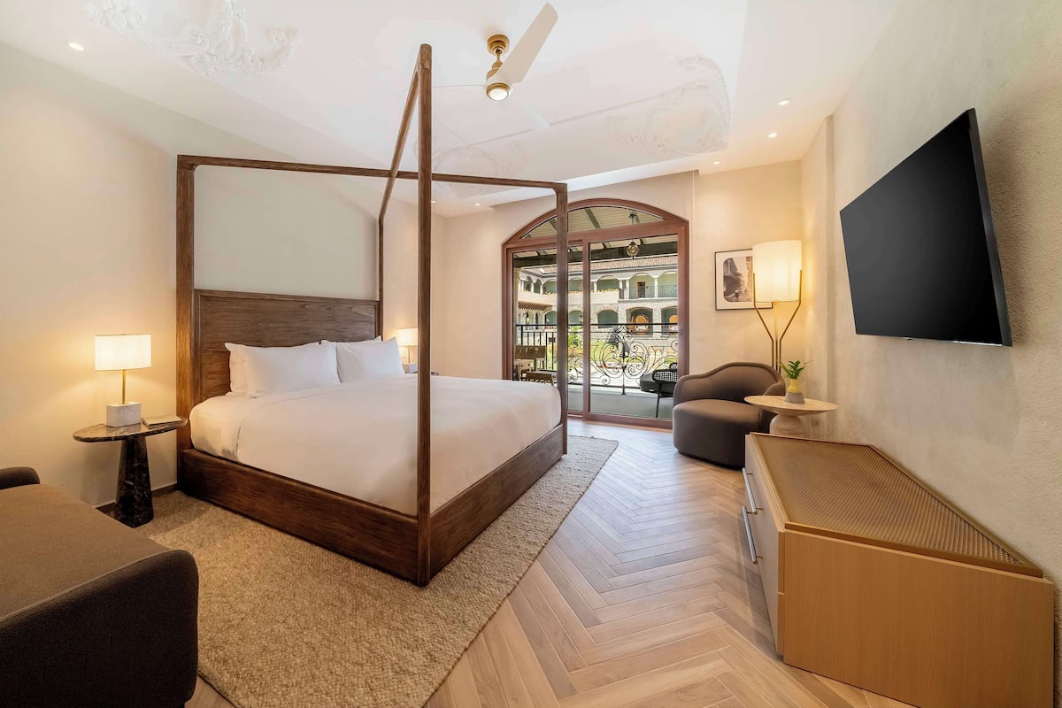 Une chambre d’hôtel moderne dotée d’un lit à baldaquin, d’un téléviseur à écran plat, d’un ventilateur de plafond et offrant une vue sur une cour intérieure grâce à une porte vitrée.