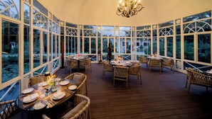 A covered outdoor dining area with tables set for two, surrounded by large windows offering views of the outdoors.