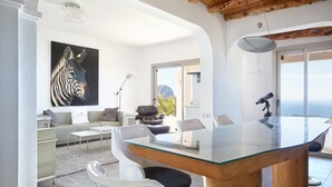 A modern living room with a glass dining table, white furniture, a zebra painting, and a view of the sea. - Beautiful Villa With sea Views (Sant Josep de sa Talaia)