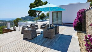 Eine Dachterrasse mit Korbmöbeln, einem blauen Sonnenschirm und Meerblick.