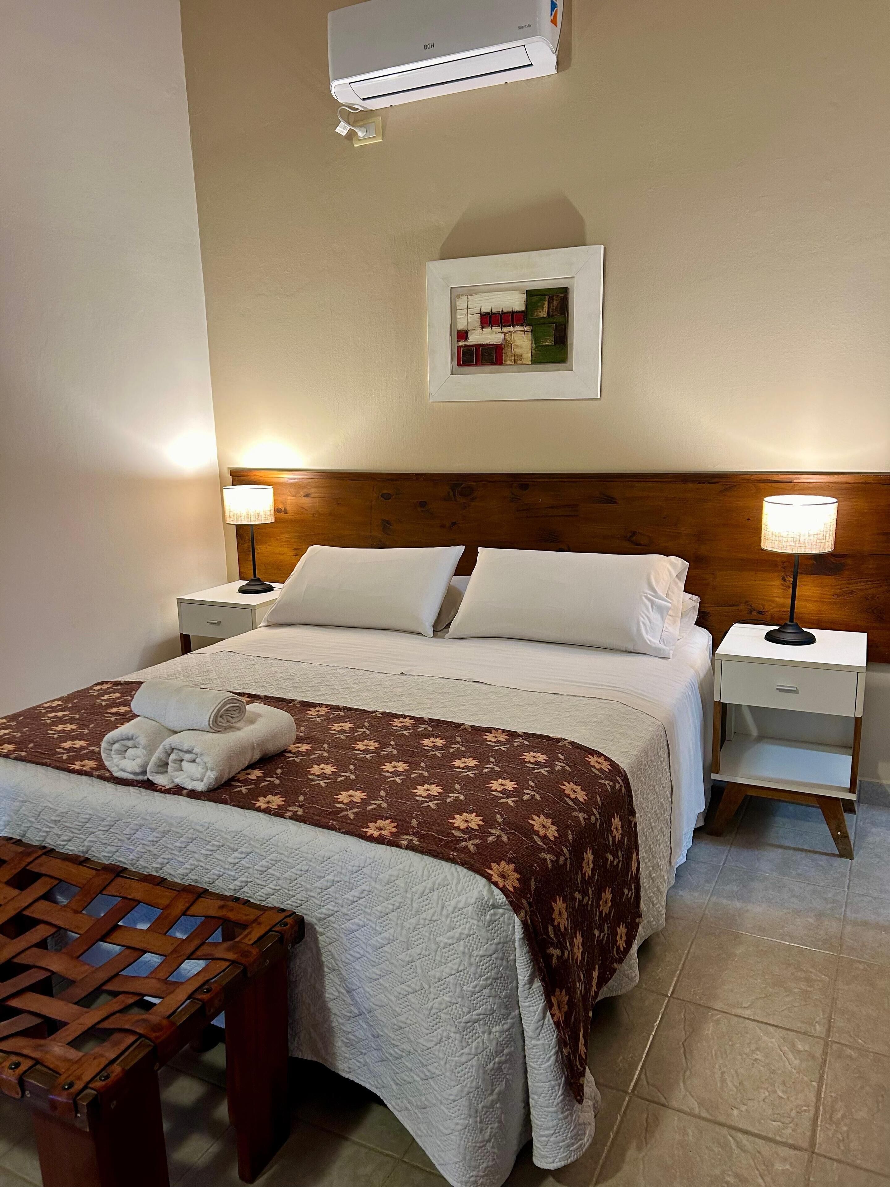 Een net opgemaakt bed met een bloemenprintdeedbed, twee bedlampjes, een ingelijst beeld op de muur en een houten bankje.