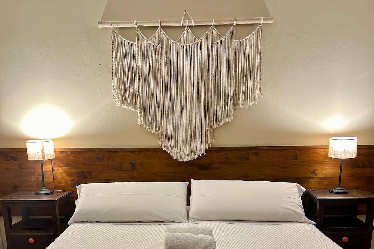 A neatly made bed with white linens, a headboard with a macrame wall hanging, two bedside lamps, and a wooden bench with a woven seat.