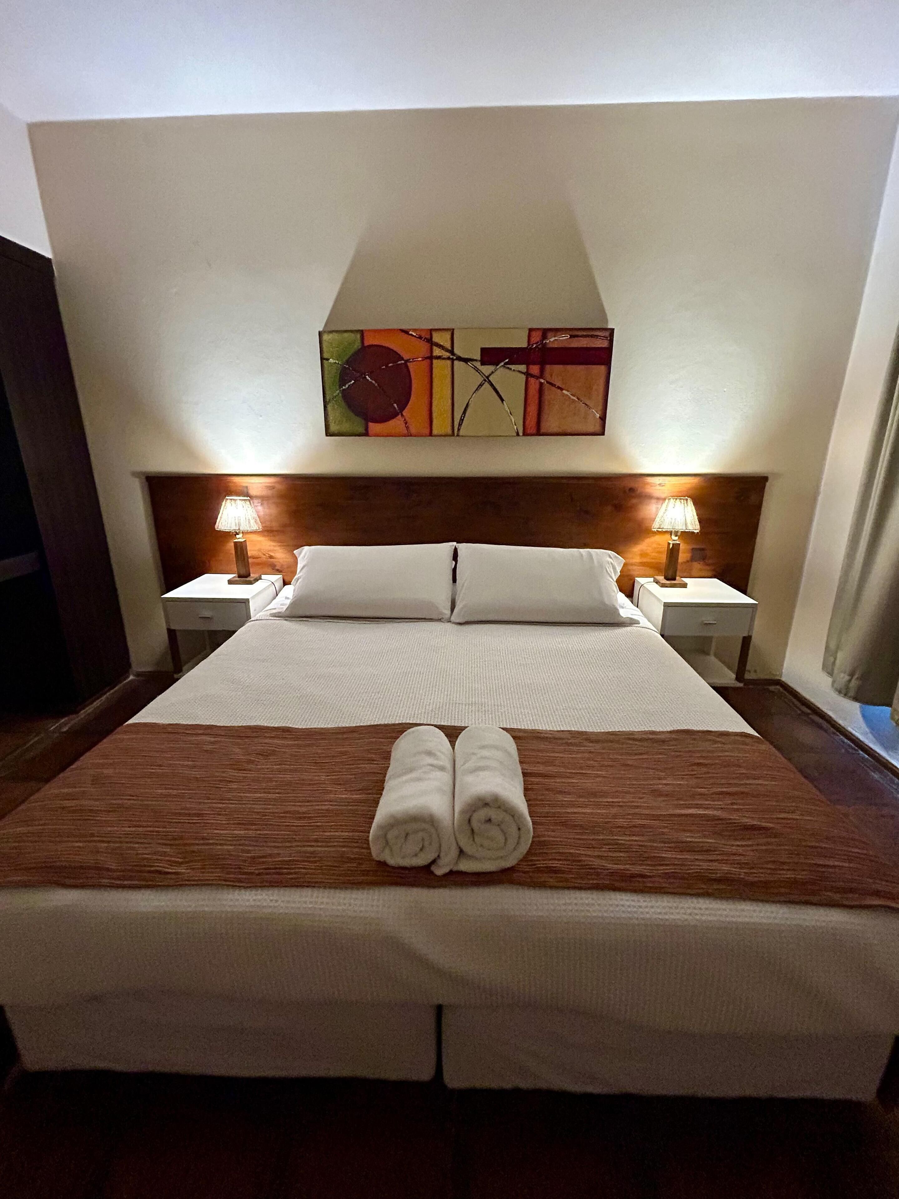 Quarto de hotel com uma cama grande, duas luminárias de cabeceira, uma cabeceira de madeira e uma pintura abstrata colorida acima da cama.
