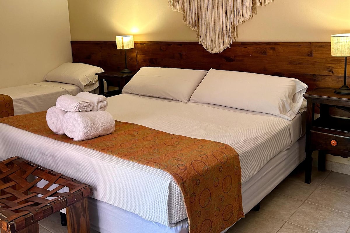 A hotel room with a large bed, two bedside tables, a wooden headboard, a macrame wall hanging, and a woven basket.