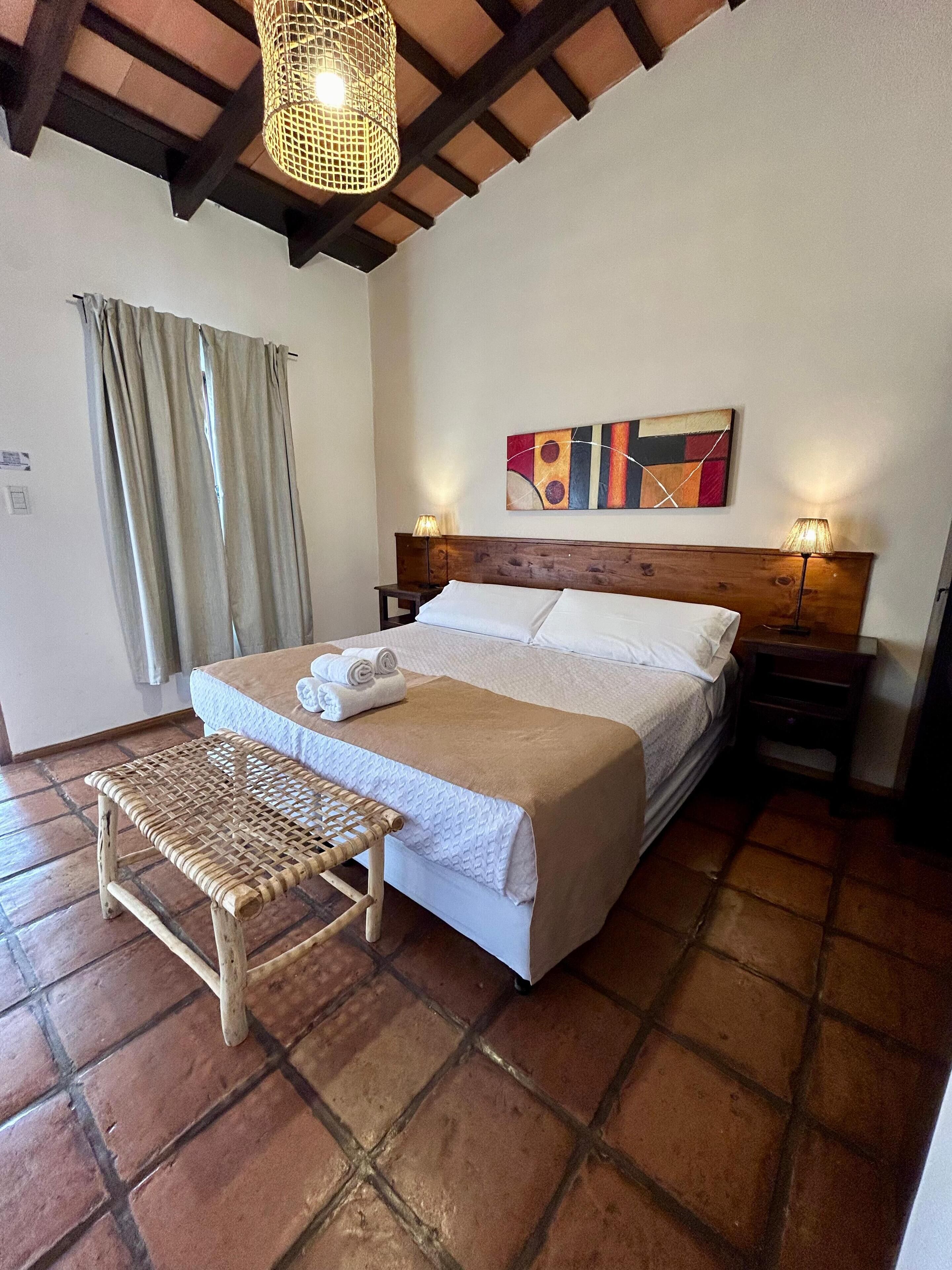 A bedroom with a large bed, wooden headboard, a woven bench, a wall painting, and a ceiling light fixture.