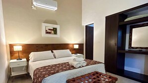 Une chambre d’hôtel avec un lit, une table de chevet, un banc en bois, un climatiseur fixé au mur et un tableau encadré accroché au mur.