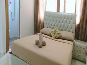 Une chambre à coucher avec une tête de lit capitonnée, un lit avec une literie beige, un ours en peluche sur le lit, une table de chevet et une porte en bois.
