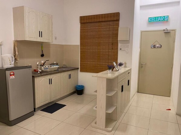 Cocina compacta con armarios blancos, refrigerador, estufa y un lavamanos pequeño. Hay una ventana con persiana de bambú, una puerta con la leyenda 'KELUAR' y un cubo de basura.