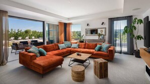 Una sala de estar moderna con un amplio sofá modular, una mesa de centro y un televisor. Amplias ventanas ofrecen vistas al exterior.