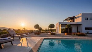 Un área moderna junto a la piscina con sillones de descanso, una alberca y un edificio contemporáneo con techo plano y ventanas amplias.