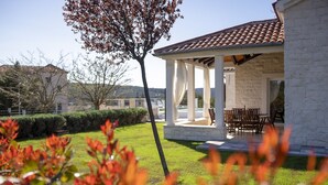 Ein überdachter Patio-Bereich mit einer weißen Steinmauer, einem Holztisch und -stühlen sowie Blick auf einen gepflegten Rasen und entfernte Gebäude.