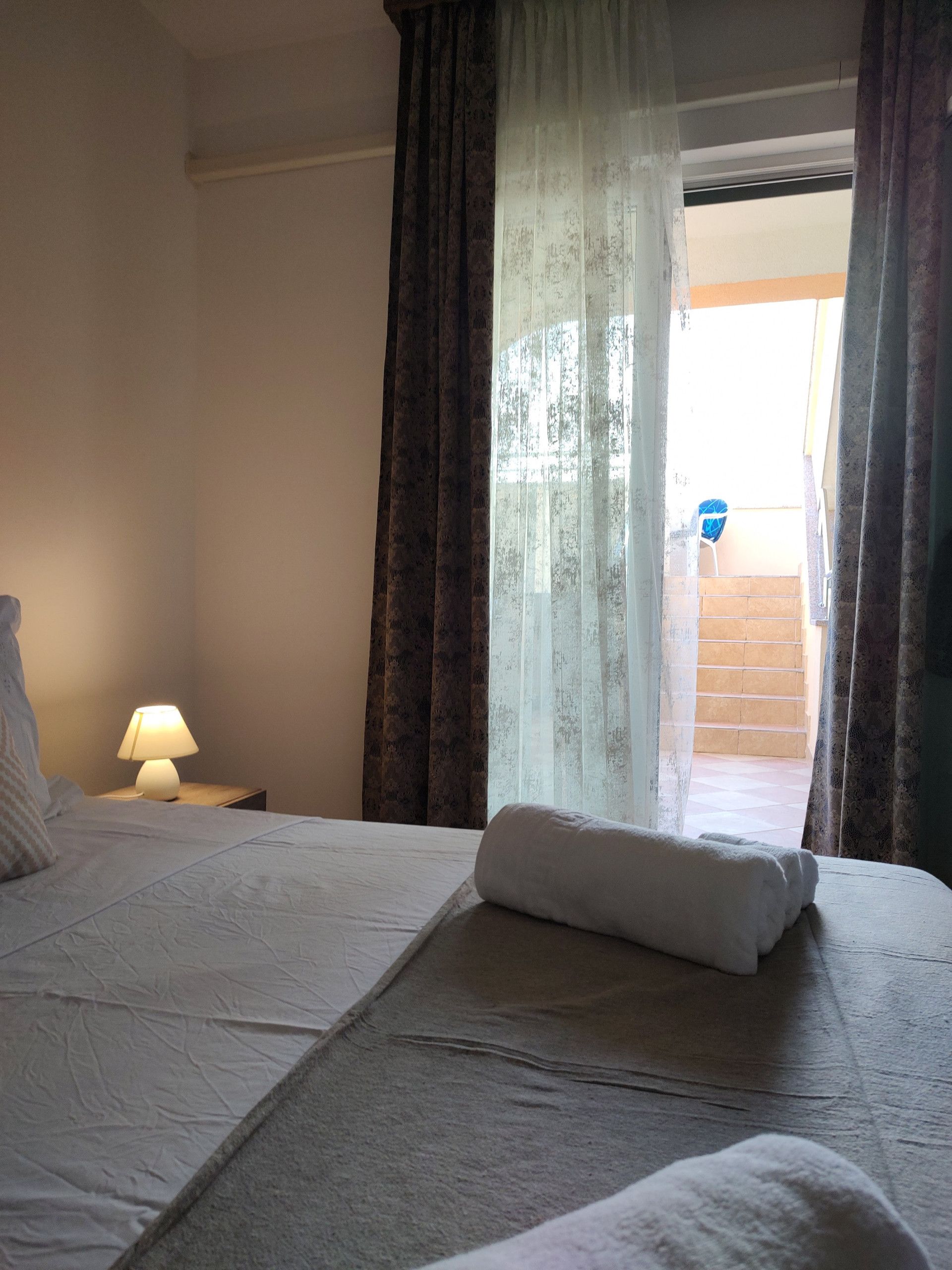 Una camera d'hotel con un letto, un comodino con una lampada e vista attraverso una finestra con tende.