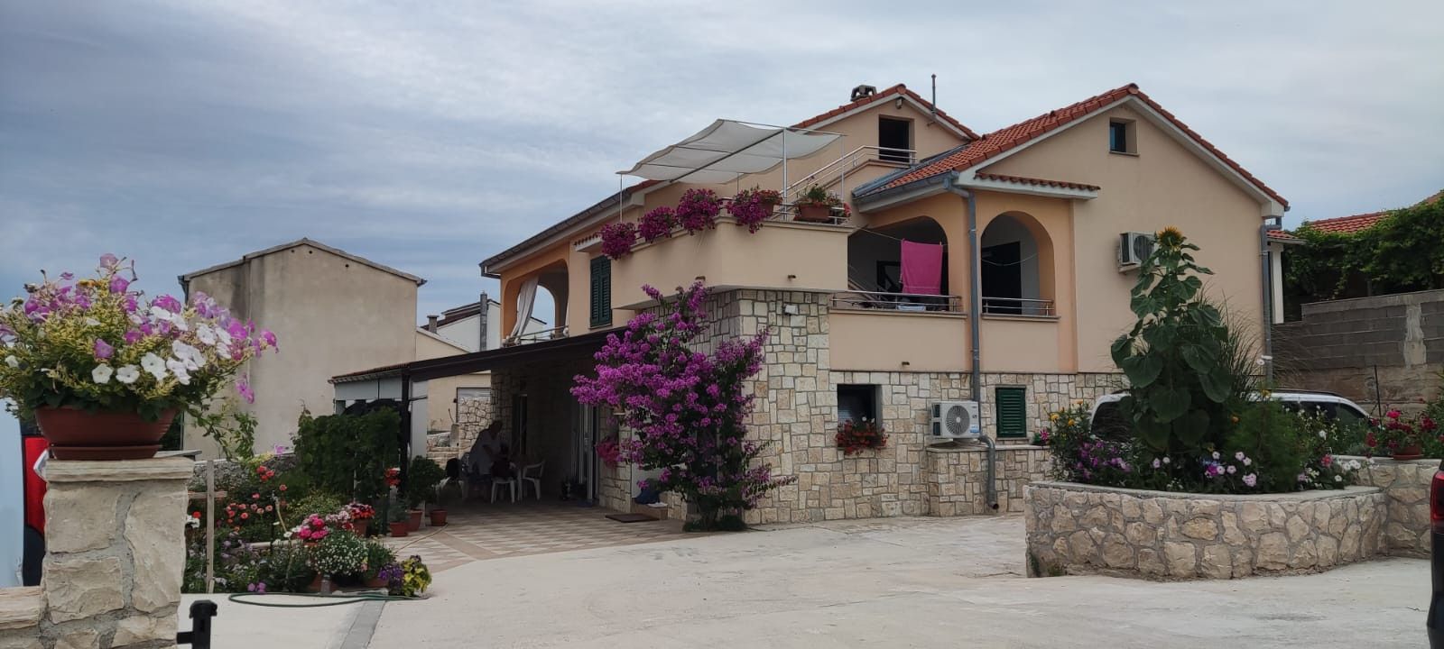 Une maison à deux étages avec une façade en pierre, un balcon orné de fleurs violettes et une voiture garée dans l’allée.