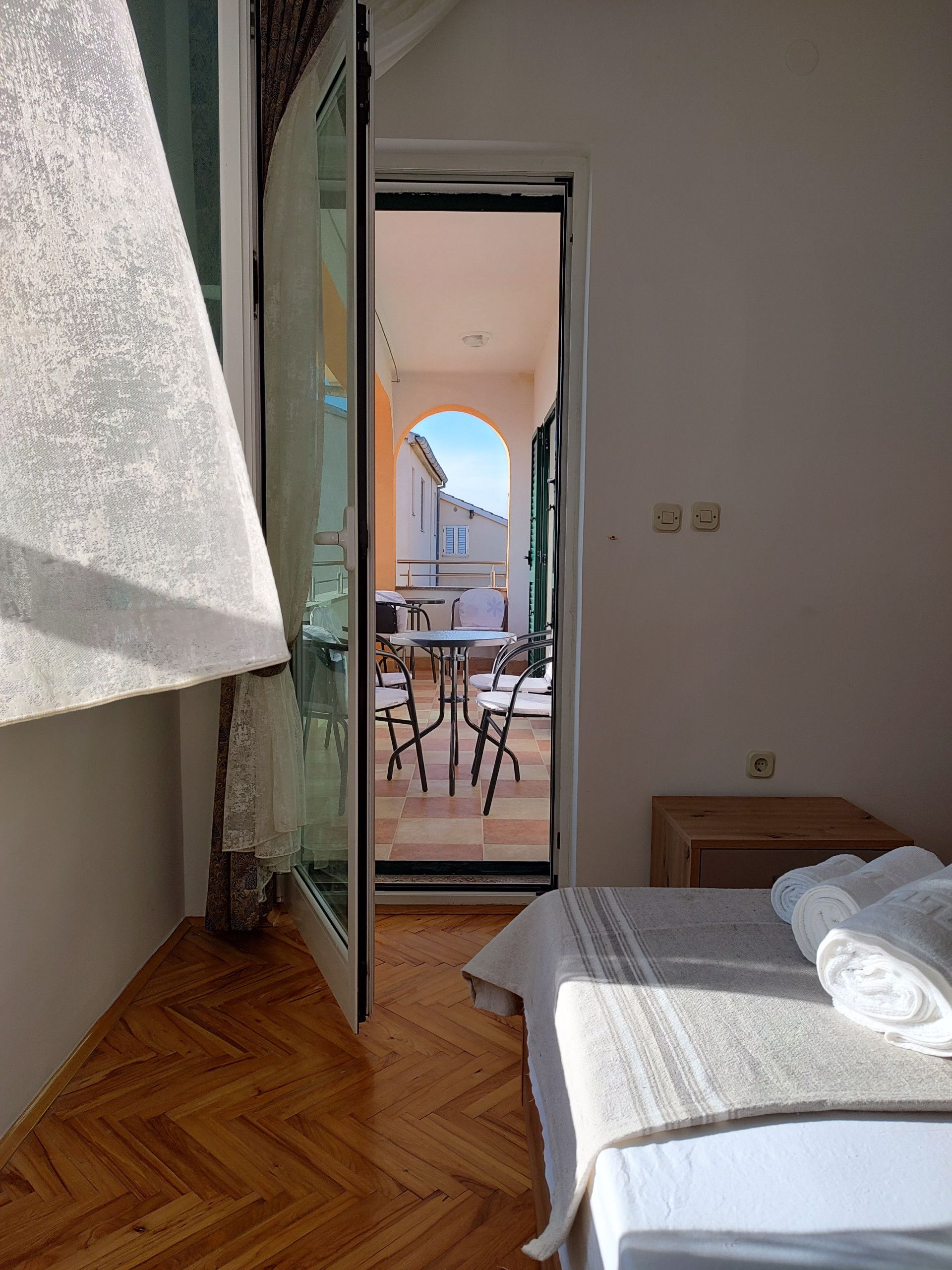 A bedroom with a wooden floor, a bed with white linens, a window with a sheer curtain, and a view of an outdoor patio with a table and chairs.