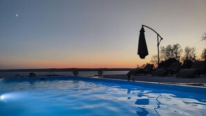 Una piscina con sillas de descanso y sombrillas, con el atardecer de fondo.