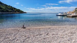 Vista despejada de una playa de guijarros con barcos amarrados a la derecha, un mar tranquilo y un paisaje montañoso al fondo.